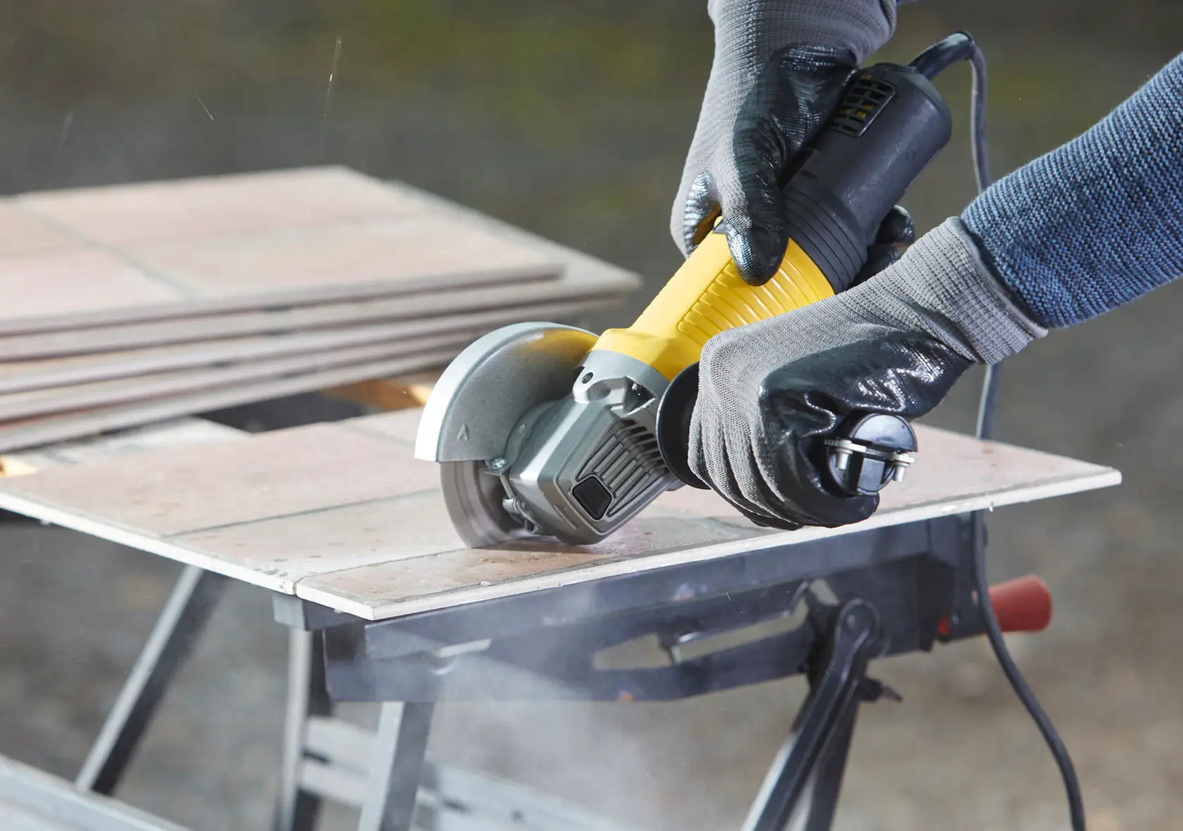 STANLEY FMEG210 angle grinder cutting tile, black and yellow body, user with gloves, close-up action shot.