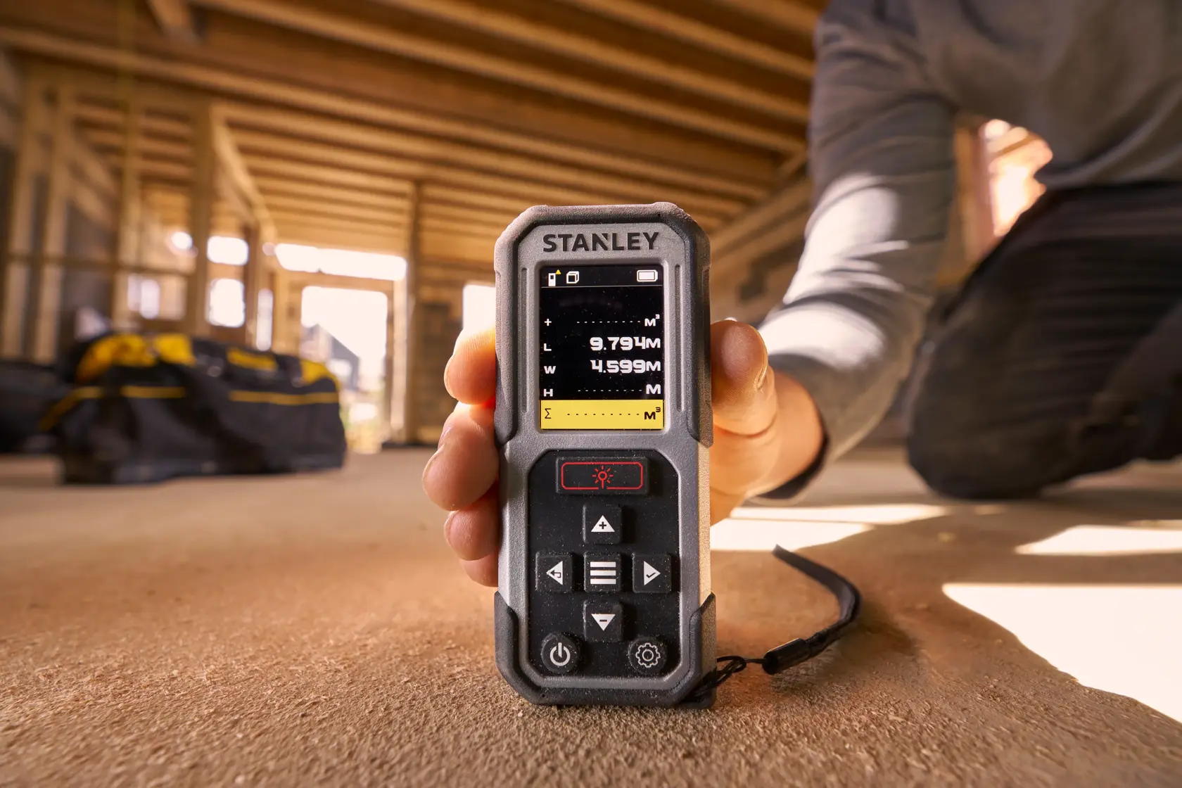 Photo of STANLEY FMHT77050-XJ laser distance measurer being held in a construction setting, display screen visible.