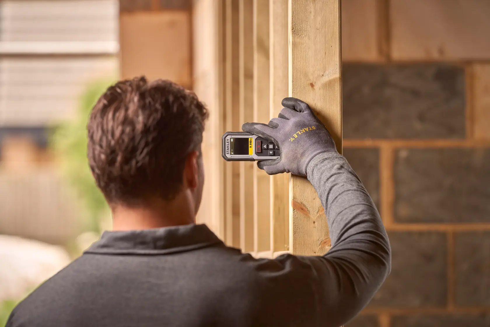 STANLEY FMHT77050-XJ laser distance measurer held by gloved hand against wooden beam, construction site background.
