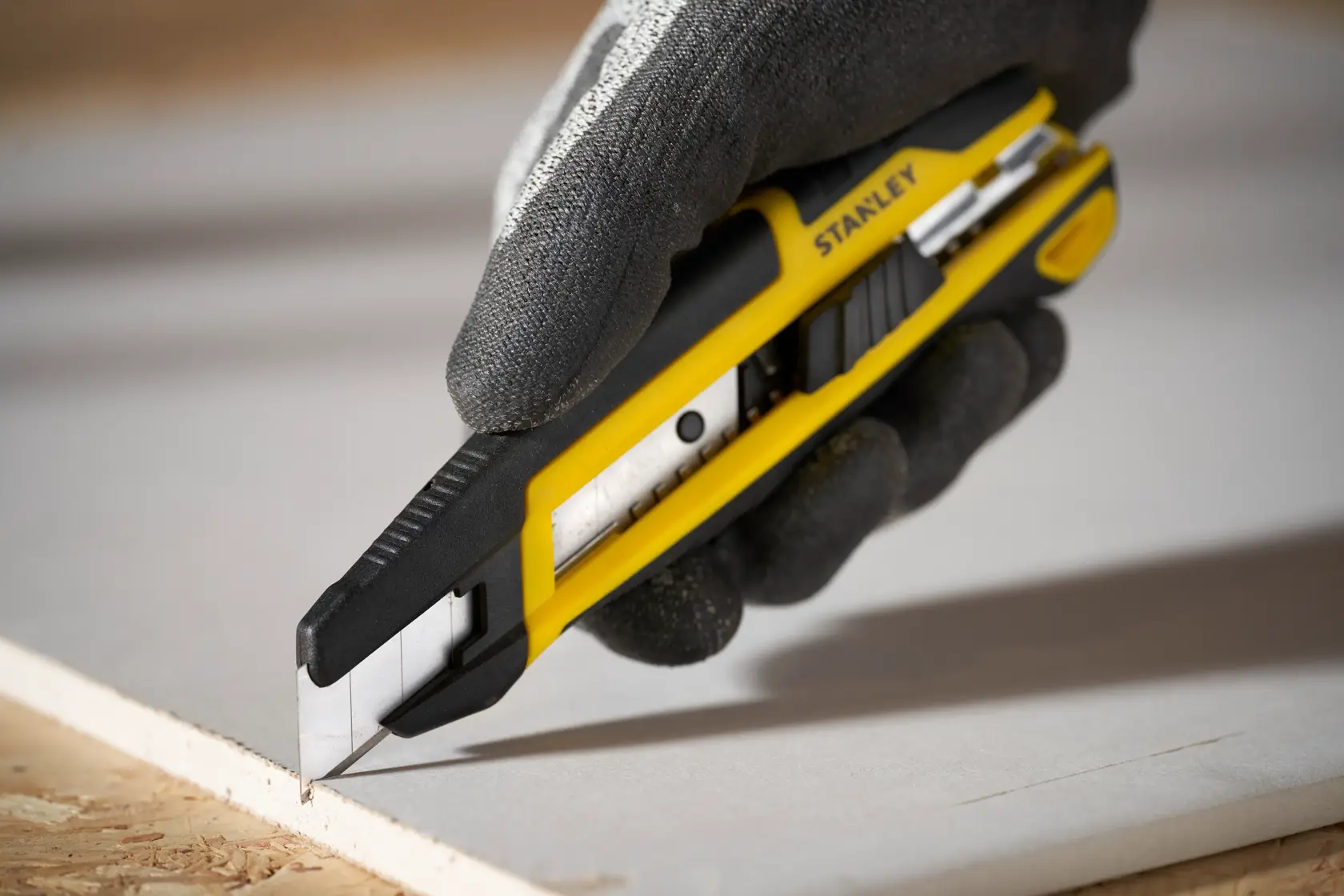 Photo of STANLEY STHT10500-0 utility knife with yellow and black body cutting drywall, held in gloved hand.