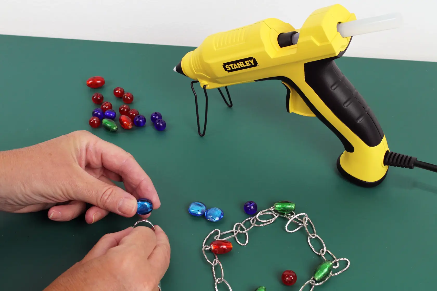 Stanley STHT6-70417 yellow glue gun for crafts shown with beads and jewelry chain on green surface.