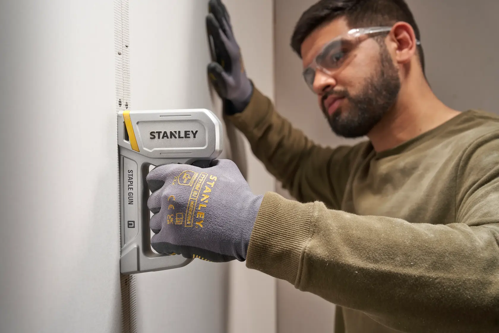 Photo of STANLEY STHT70150-0 staple gun used on wall, person wearing STANLEY gloves holding the tool.