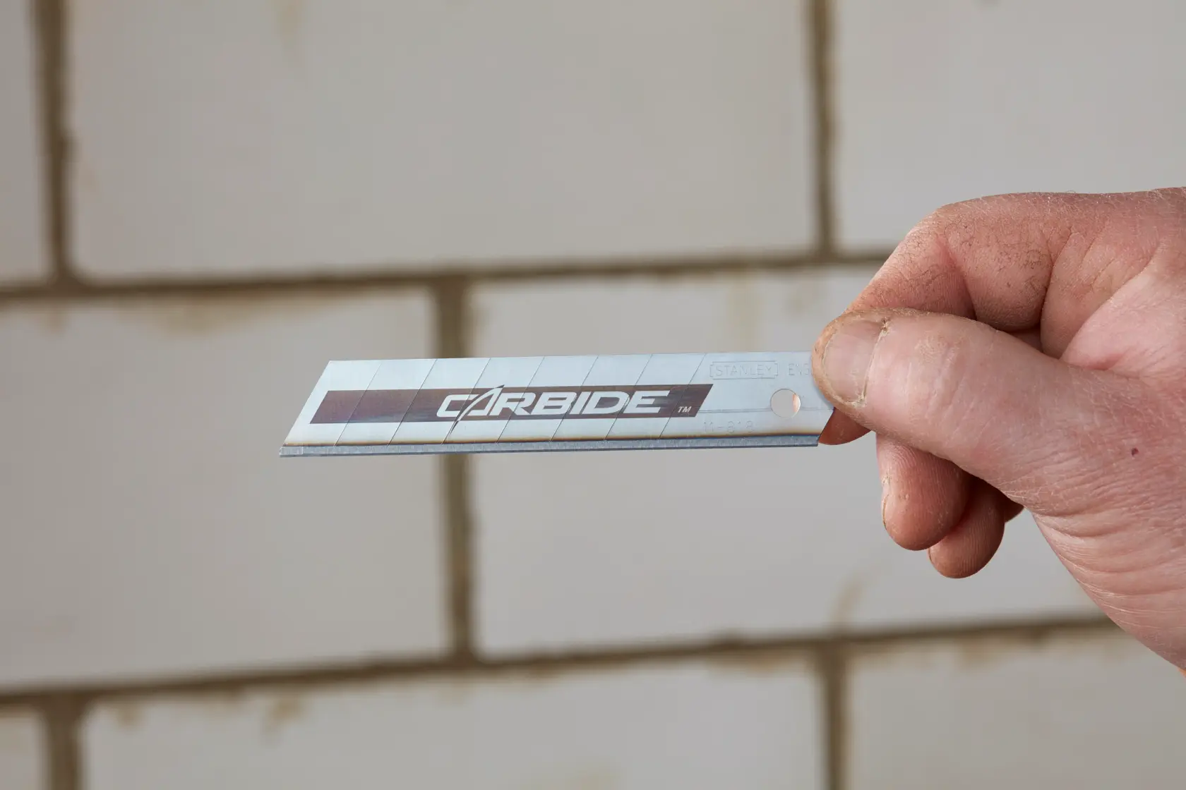 Photo of STANLEY STHT8-11818 carbide utility blade held in hand with tiled wall background.