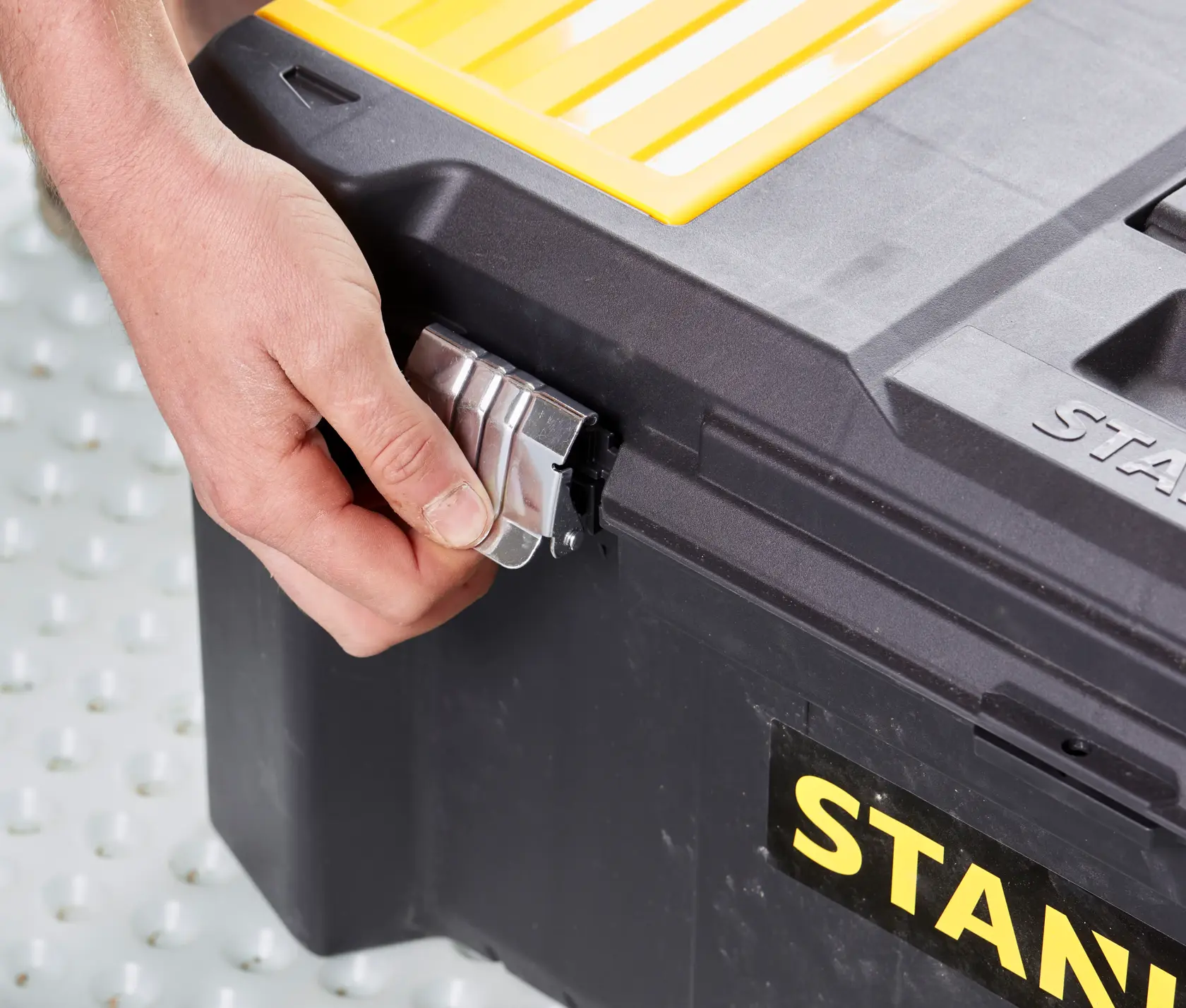 STANLEY STST82976-1 toolbox close-up, hand unlocking metal latch on corner, black and yellow plastic case.